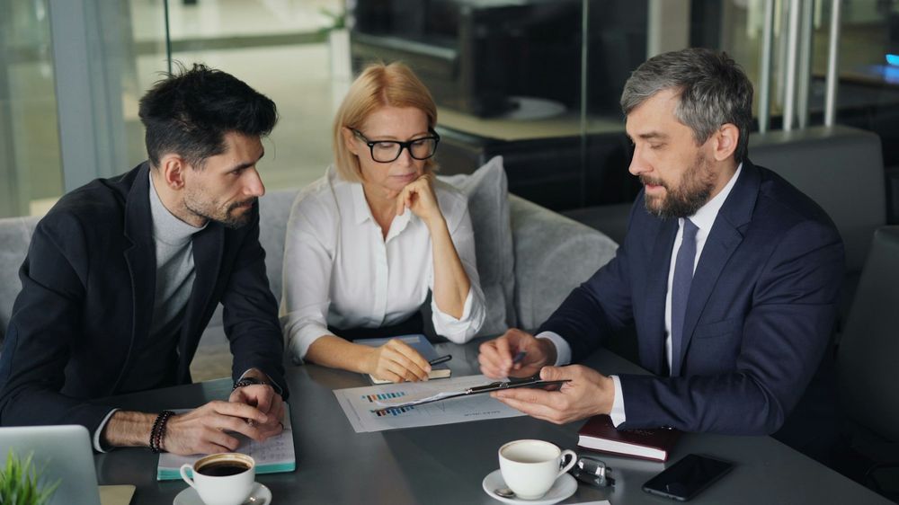 Três profissionais de terno analisando relatórios em uma mesa de reunião